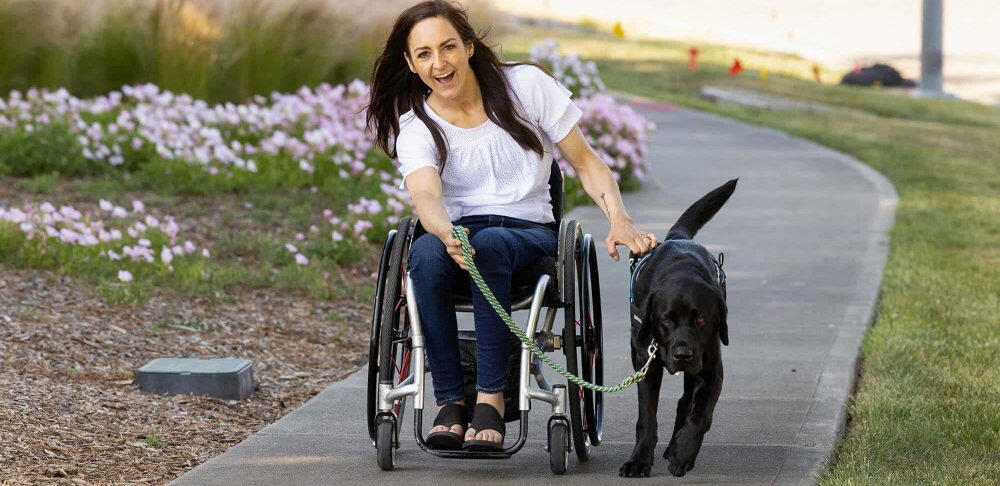 Una mujer en silla de ruedas, sonriendo, es guiada por un perro de asistencia Labrador negro en un camino pavimentado, rodeado de césped y flores rosadas.